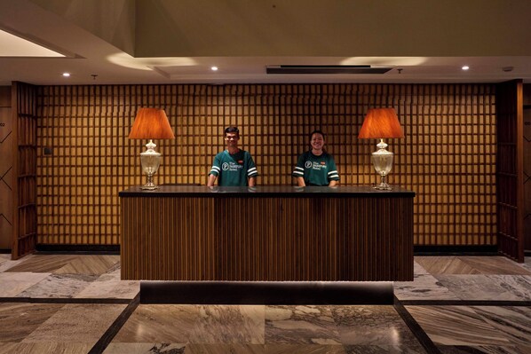 A reception desk with two staff members, wooden paneling on the wall, and two lamps. - The Passport Hotel (Vagator)