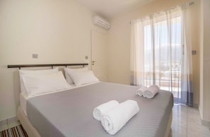 A hotel room with a bed, a window with sheer curtains, and an air conditioning unit on the wall.