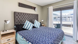 A bedroom with a large bed, a headboard, two bedside lamps, a wooden nightstand, and a view of a balcony with a railing. - Seaside Village J9 by CTHA (Cape Town)