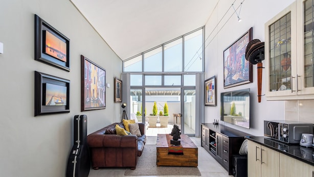 A modern living room with a sofa, coffee table, and guitar, leading to a kitchen with a large window and artwork on the walls. - Seaside Village J9 by CTHA (Cape Town)