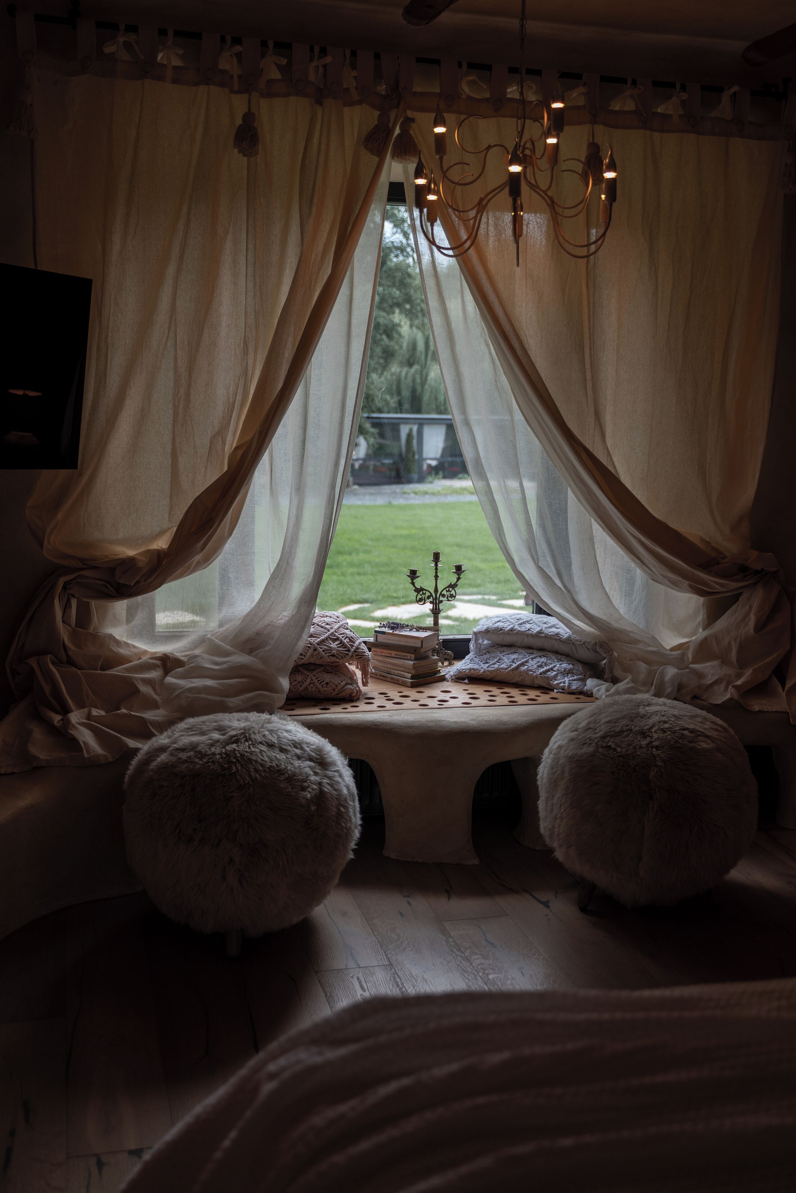 Una habitación acogedora con un ventanal grande, cortinas traslúcidas, una araña y una pequeña mesa con libros y almohadones.