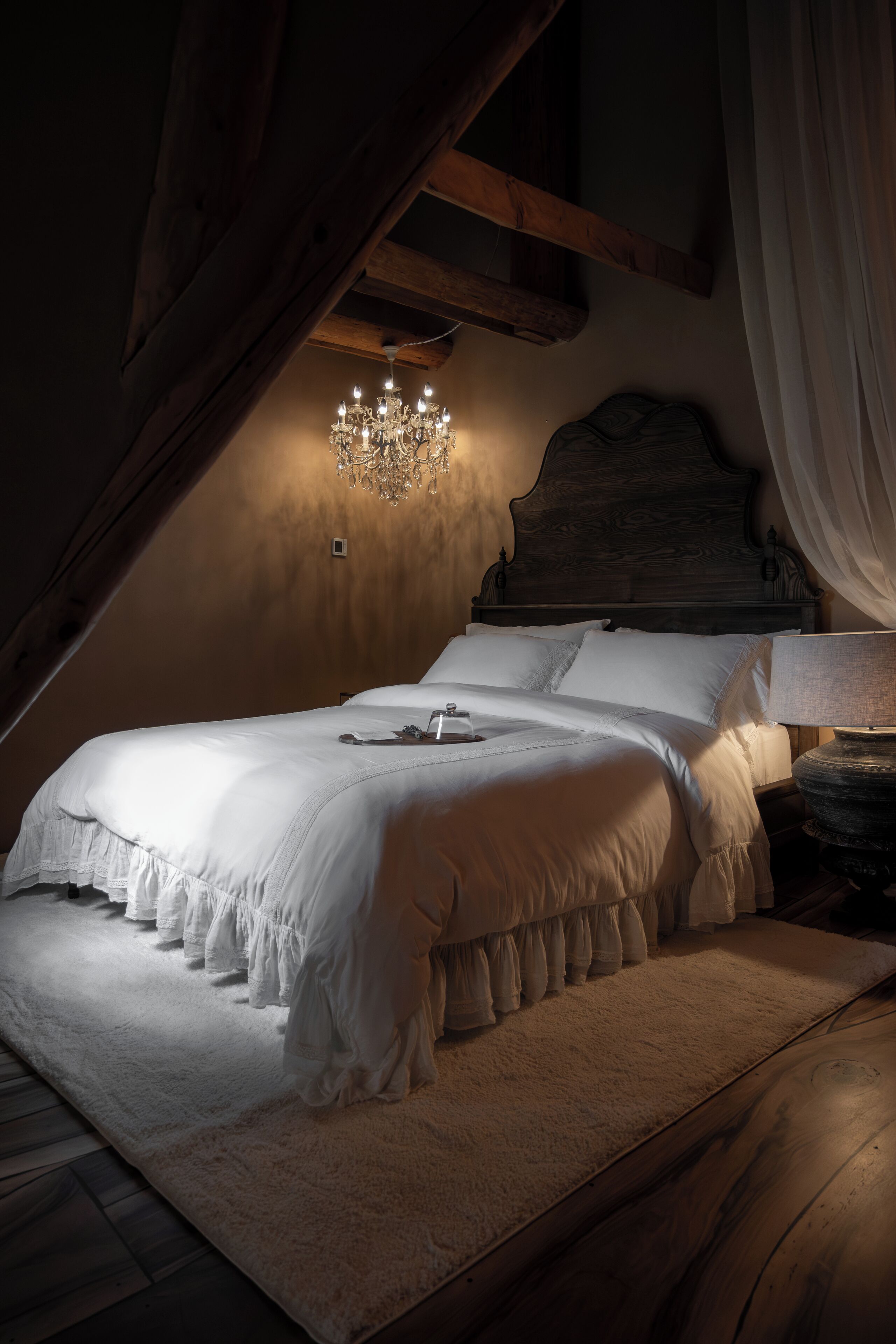 A bedroom with a wooden ceiling, a chandelier, a bed with white bedding, and a nightstand with a lamp.