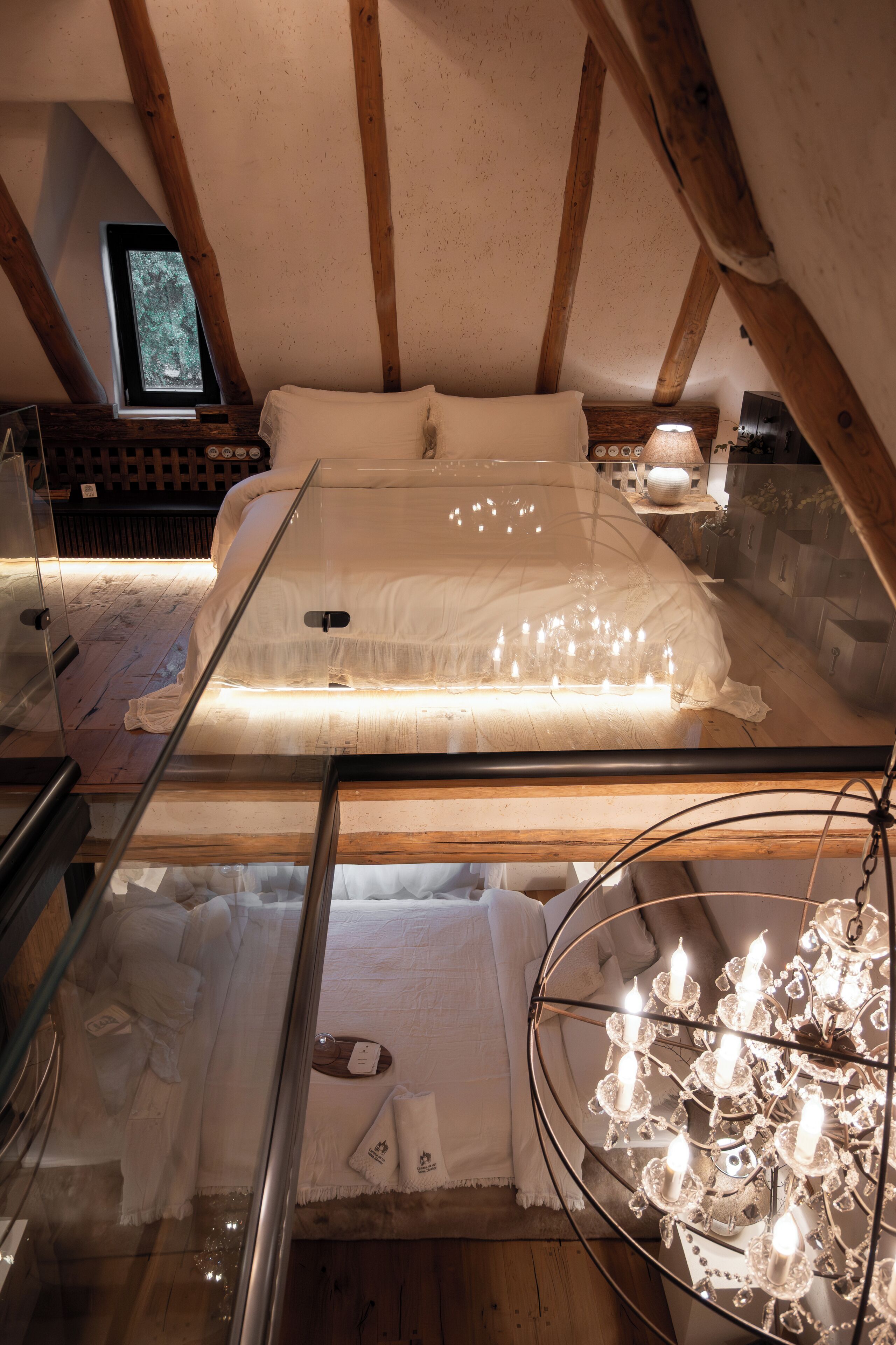 Un dormitorio con una cama de cristal, vigas de madera, una araña y una mesita de noche con lámpara.