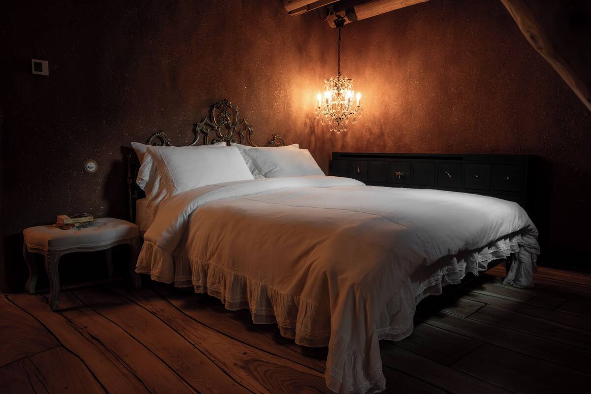 A bedroom with a wooden floor, a bed with white linens, a nightstand, and a wall-mounted chandelier.