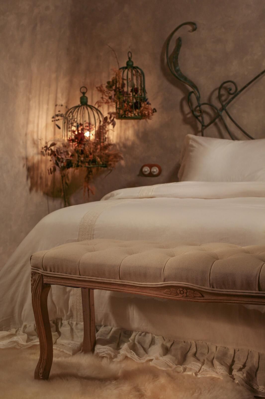 A bed with a tufted headboard, a bedside table with a vase of dried flowers, and a wall-mounted decorative metal piece.