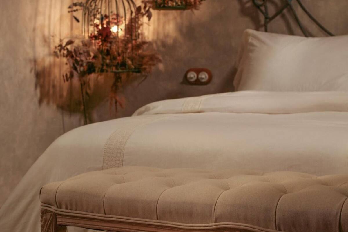 A bed with a tufted headboard, a bedside table with a vase of dried flowers, and a wall-mounted decorative metal piece.