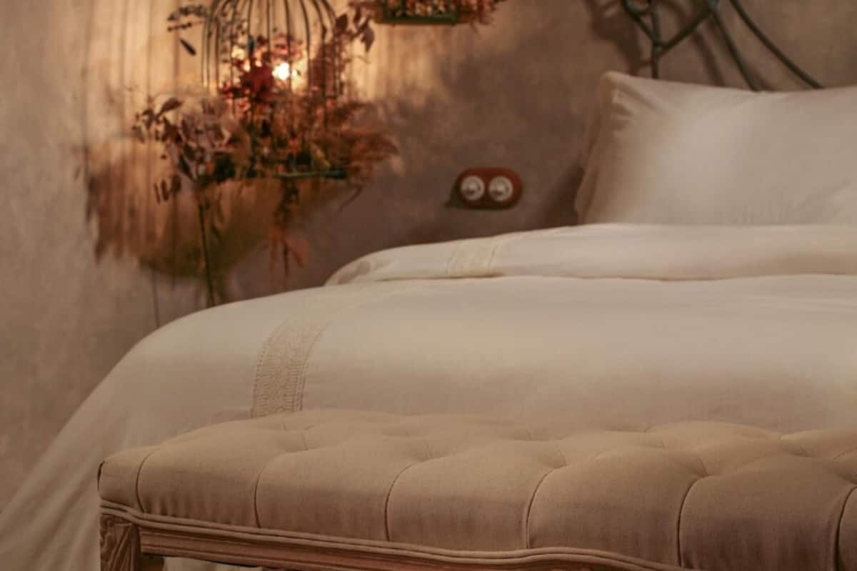 A bed with a tufted headboard, a bedside table with a vase of dried flowers, and a wall-mounted decorative metal piece.