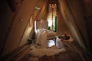 Un dormitorio cálido en el ático, con piso de madera, una ventana grande con cortina, un candelabro y una alfombra de piel.