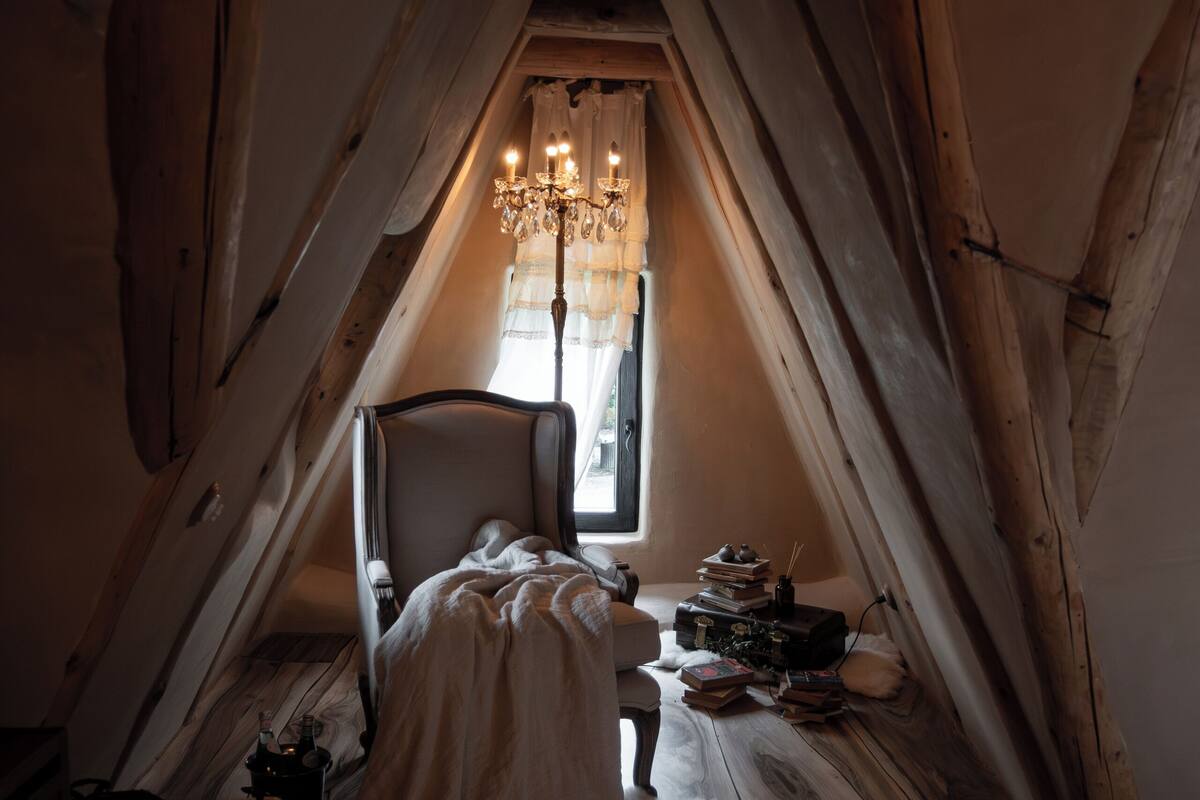 A narrow attic bedroom with a sloped ceiling, a bed, a chair, a dresser, and a chandelier.