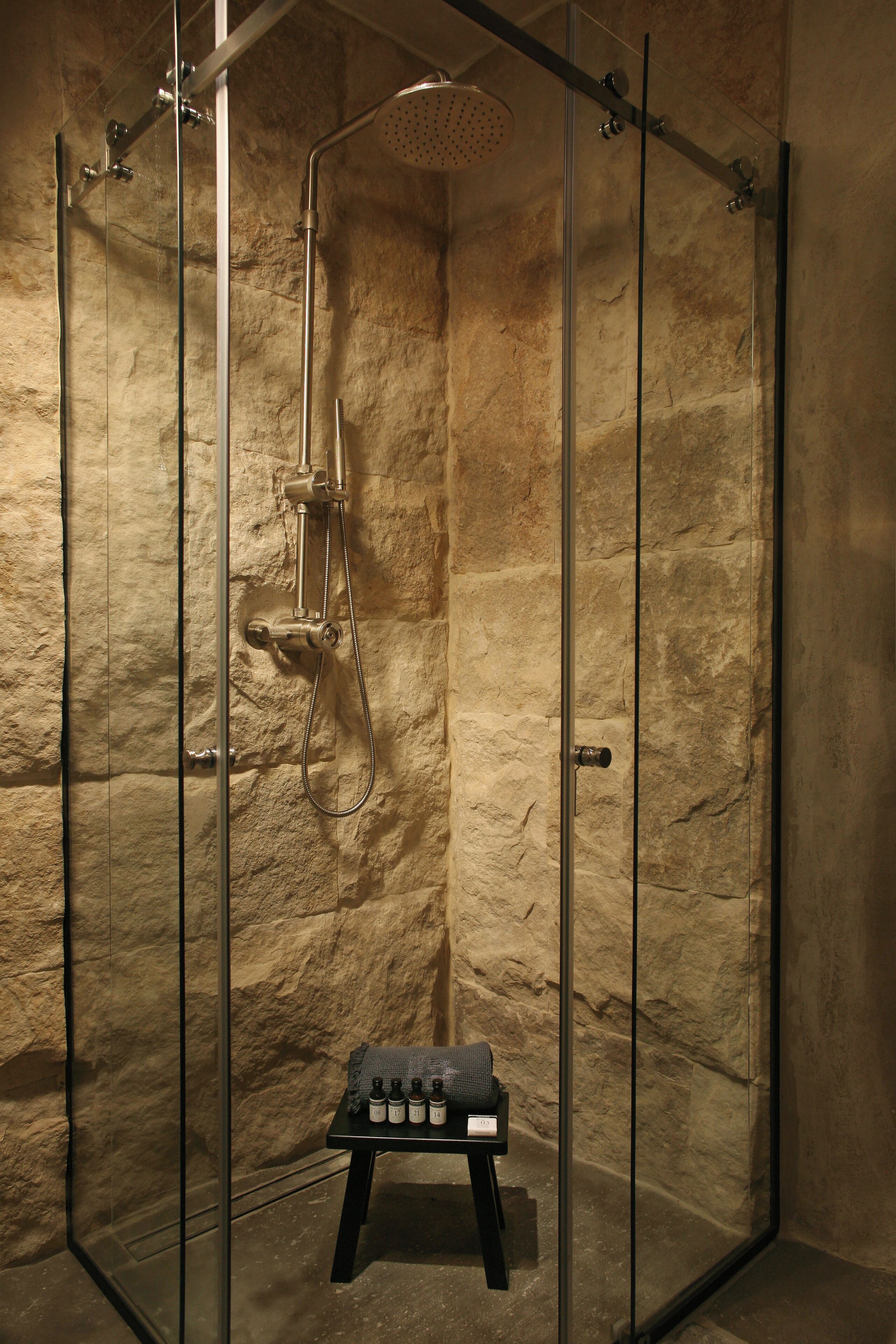 Eine gläserne Dusche mit Wand aus Stein, einer Handbrause und einem kleinen Tisch mit Flaschen.