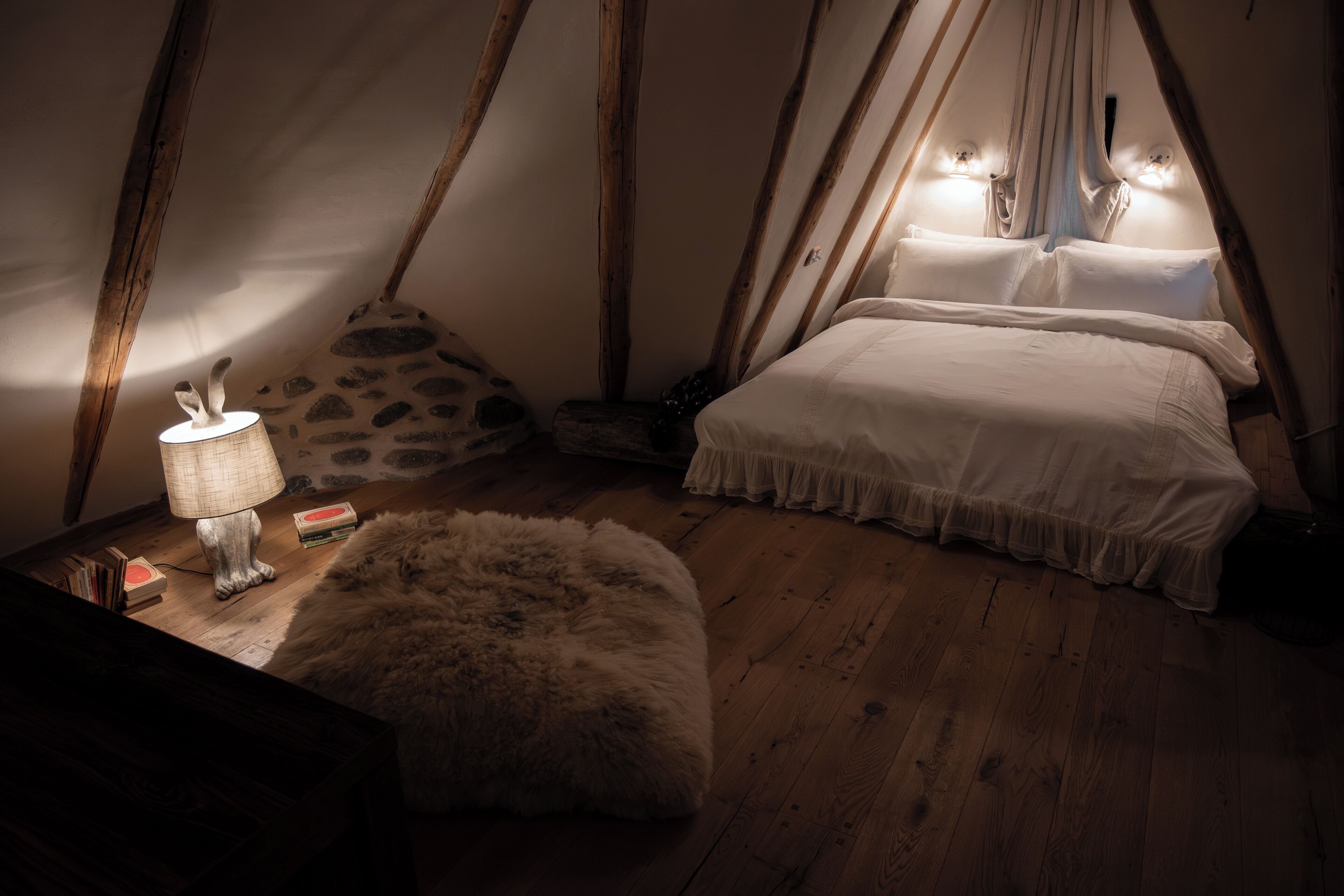 Una camera da letto accogliente e scarsamente illuminata, con pavimento in legno, un pouf in pelliccia e un letto con lenzuola bianche, più una lampada su un comodino.