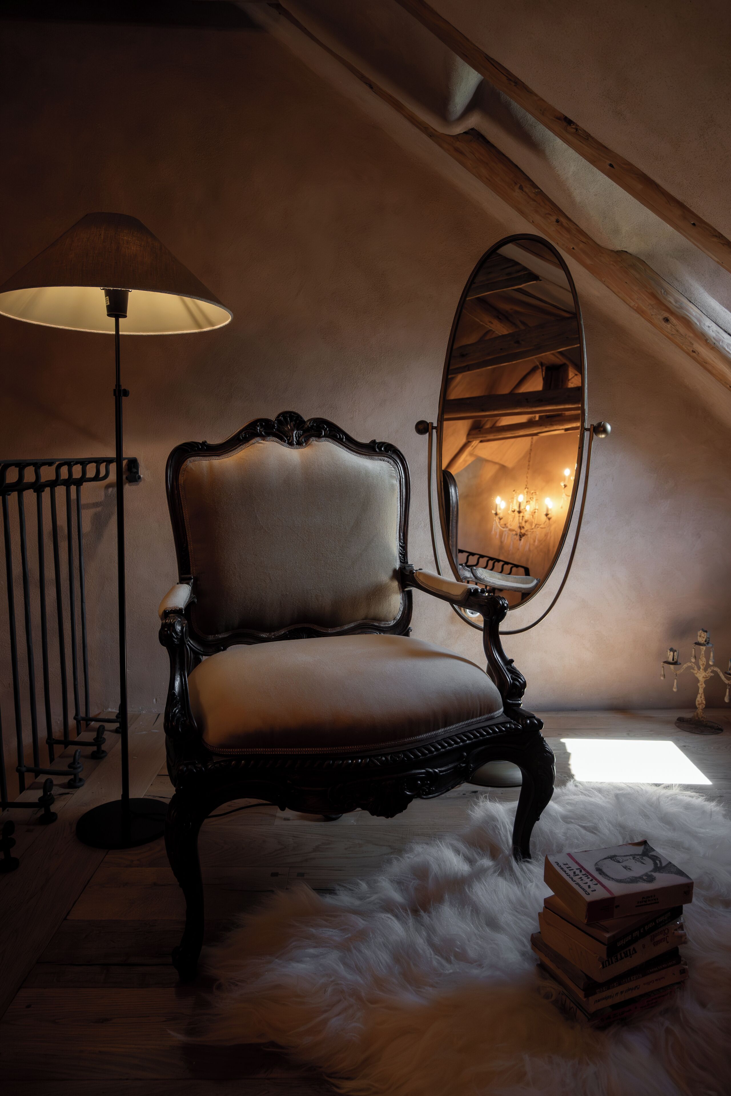 A chair with a mirror, a lamp, and a stack of books in a room with a sloped ceiling and wooden beams.