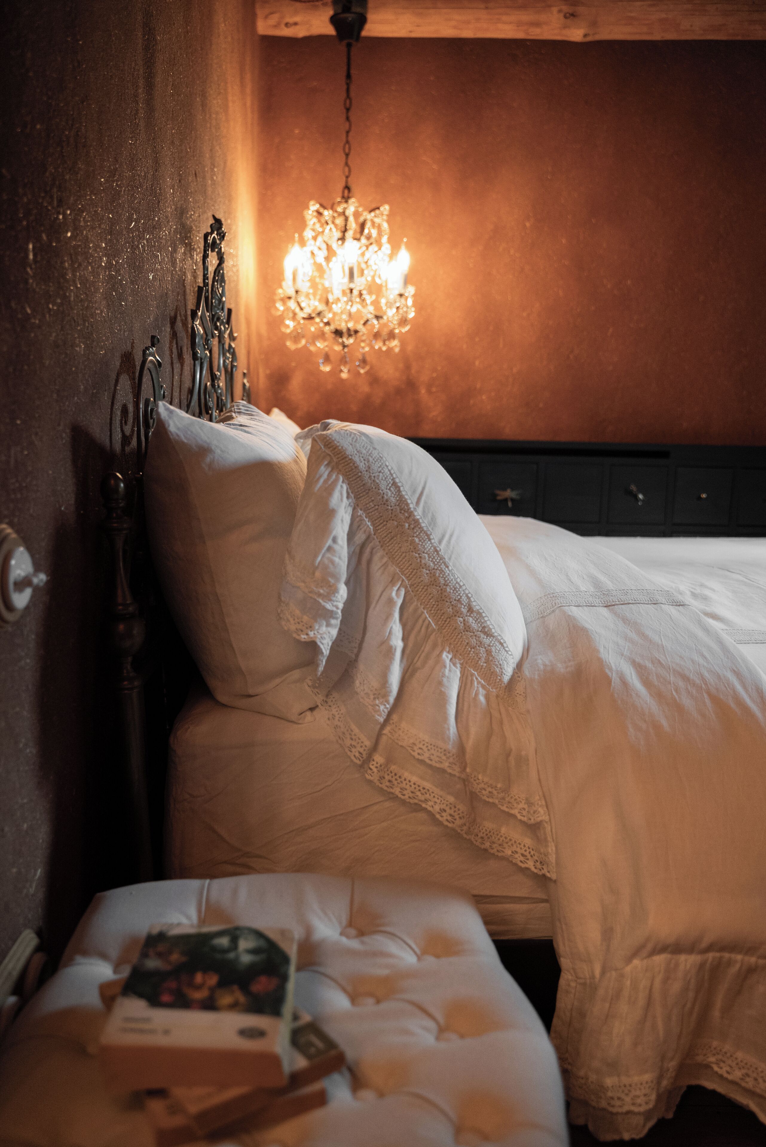 A neatly made bed with a white comforter and lace-trimmed pillows, a bedside table with a book, and a chandelier hanging above.