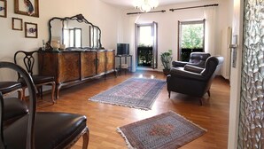Amplia sala de estar con piso de madera, sofá de cuero negro, aparador de madera y un espejo grande. Hay una araña colgando del techo y una ventana con cortinas.