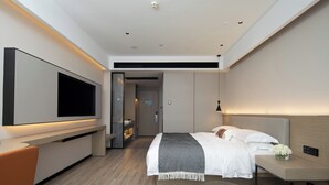 A modern hotel room with a large flat-screen TV, a bed with white bedding, a bedside table with a vase of flowers, and a glass coffee table with toiletries. - Yishang Hotel  (Chengmai)