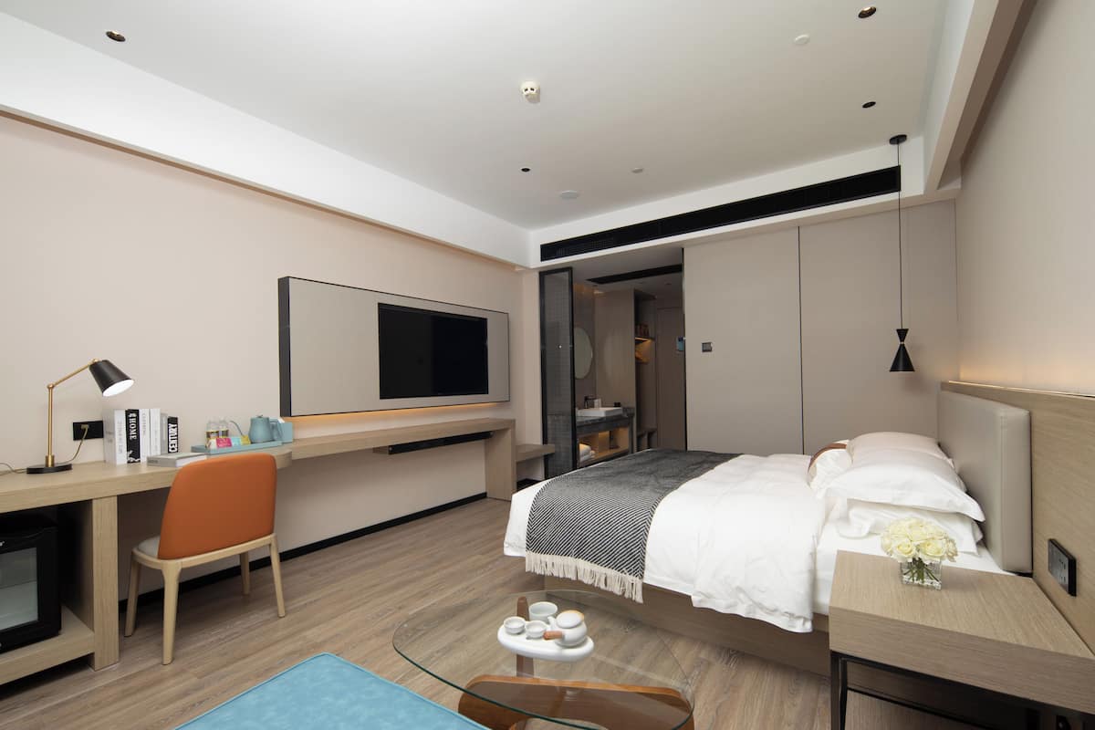 A modern hotel room with a bed, a desk, a chair, a TV, and a bedside table with a vase of flowers.