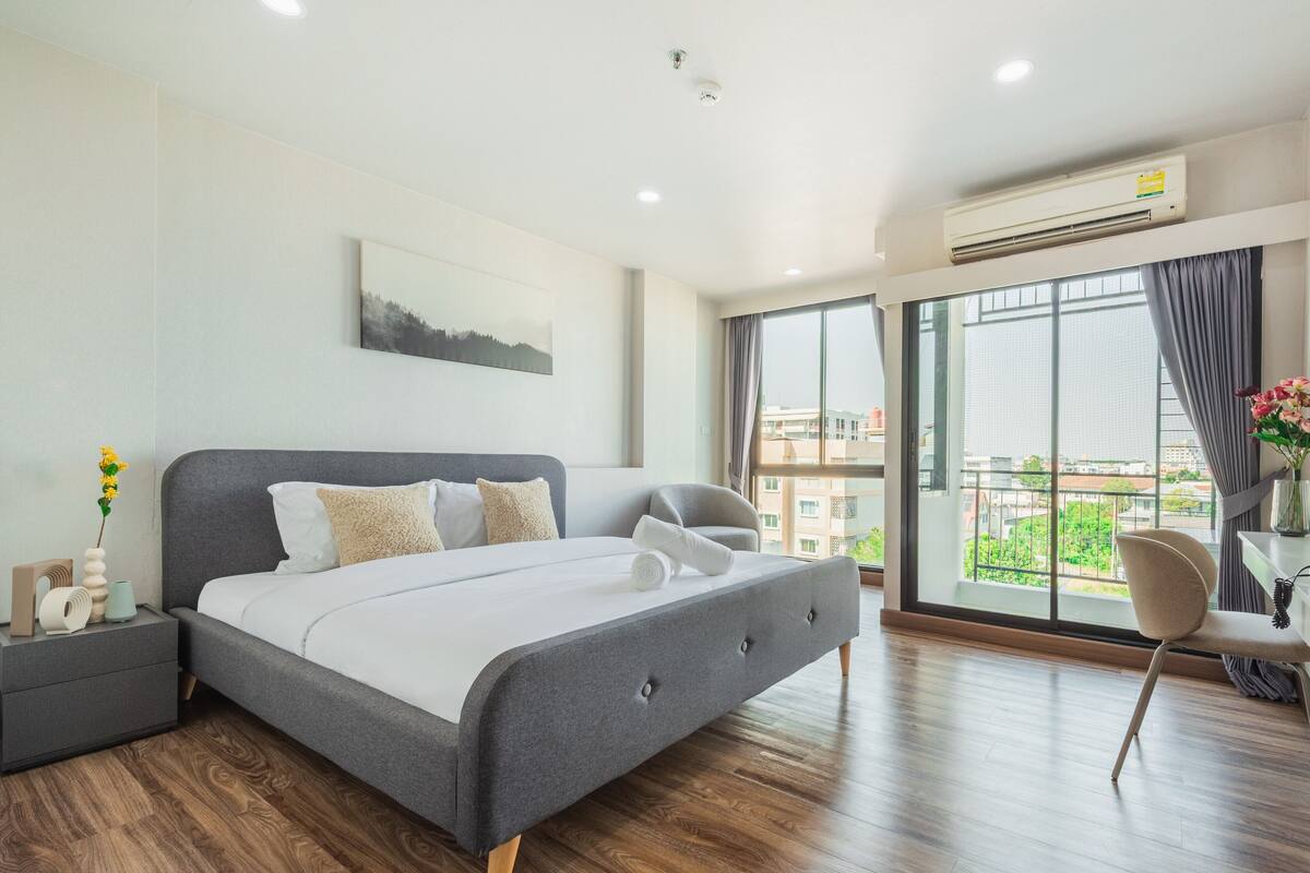 Ein modernes Schlafzimmer mit einem großen Bett, einem Nachttisch, einem Sessel und Blick auf die Stadt durch große Fenster.
