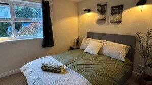 A bedroom with a bed, a window with curtains, a wall-mounted lamp, and a small plant. - Winsford Woodford Cottage - Sleeps 6 - Pets Ok (Winsford)