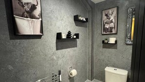 A bathroom with a grey textured wall, a toilet, a towel rack, and two black shelves with items on them. - Winsford Woodford Cottage - Sleeps 6 - Pets Ok (Winsford)