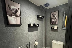 Un bagno con una parete grigia e strutturata, un WC, un portasciuga e due mensole nere con vari oggetti.