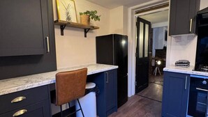 A compact kitchen with dark cabinets, a marble countertop, a single chair, and a black refrigerator. - Winsford Woodford Cottage - Sleeps 6 - Pets Ok (Winsford)