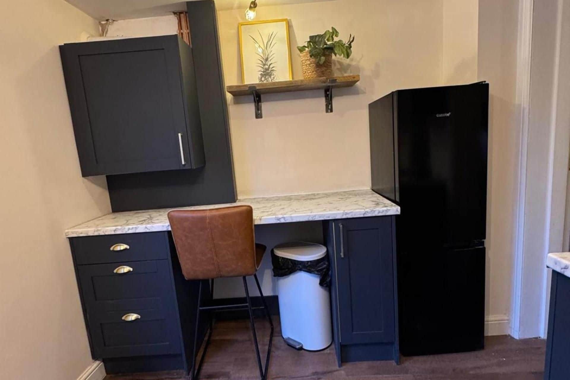 Cocina compacta con armarios oscuros, encimera de mármol, refrigerador negro, contenedor de basura y una silla marrón.