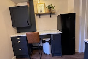 A compact kitchen with dark cabinetry, a marble countertop, a black refrigerator, a trash bin, and a brown chair. - Winsford Woodford Cottage - Sleeps 6 - Pets Ok (Winsford)