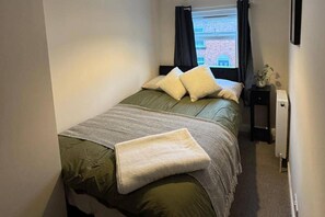 A single bed with a green bedspread and a white throw blanket, a bedside table with a lamp, and a window with curtains. - Winsford Woodford Cottage - Sleeps 6 - Pets Ok (Winsford)