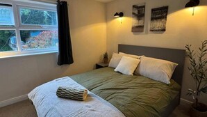 A bedroom with a bed, a window, a nightstand, a lamp, and a wall-mounted poster. - Winsford Woodford Cottage - Sleeps 6 - Pets Ok (Winsford)