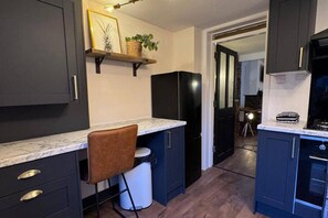 A kitchen with dark cabinets, a marble countertop, a black refrigerator, and a chair. - Winsford Woodford Cottage - Sleeps 6 - Pets Ok (Winsford)
