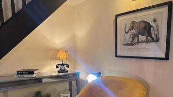 A room with a staircase, a lamp, a chair, a framed picture, and a small table with books.
