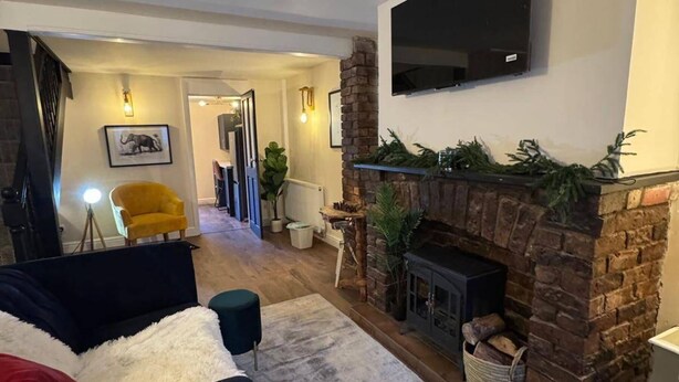 A living room with a fireplace, a sofa, a yellow armchair, and a TV mounted on the wall. - Winsford Woodford Cottage - Sleeps 6 - Pets Ok (Winsford)