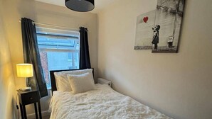 A bedroom with a bed, a bedside table with a lamp, a radiator, a window with curtains, and a wall with a piece of art. - Winsford Woodford Cottage - Sleeps 6 - Pets Ok (Winsford)