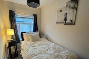 Una camera da letto con un letto, un comodino con una lampada, un termosifone, una finestra con tende e un quadro appeso al muro.