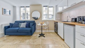 A compact living space with a blue sofa, a small round table with a drink, a washing machine, and a compact kitchen area. - Atelier d'artiste a Saint Germain (Paris)