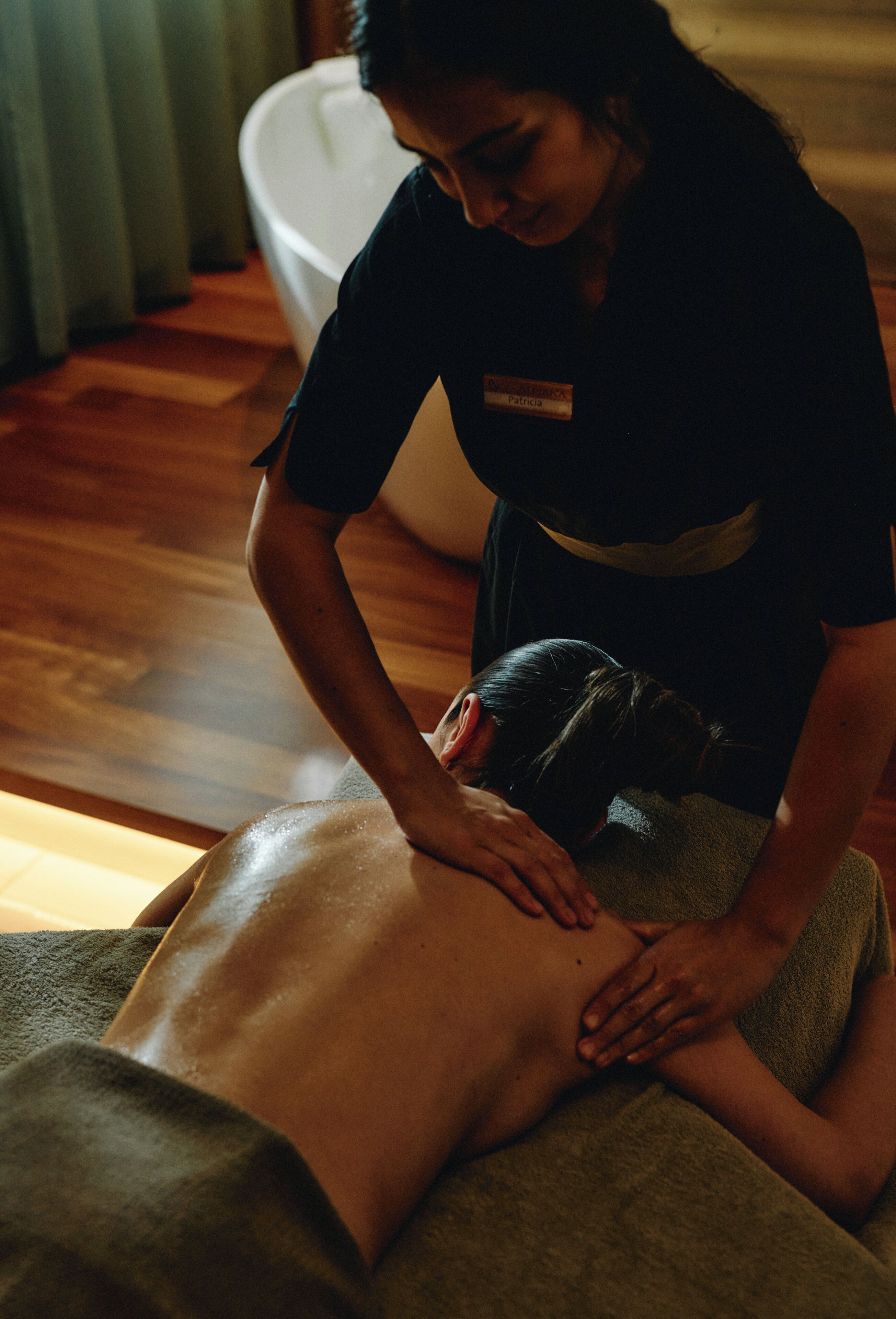 A person receiving a back massage in a room with a wooden floor and a large white bathtub in the background.