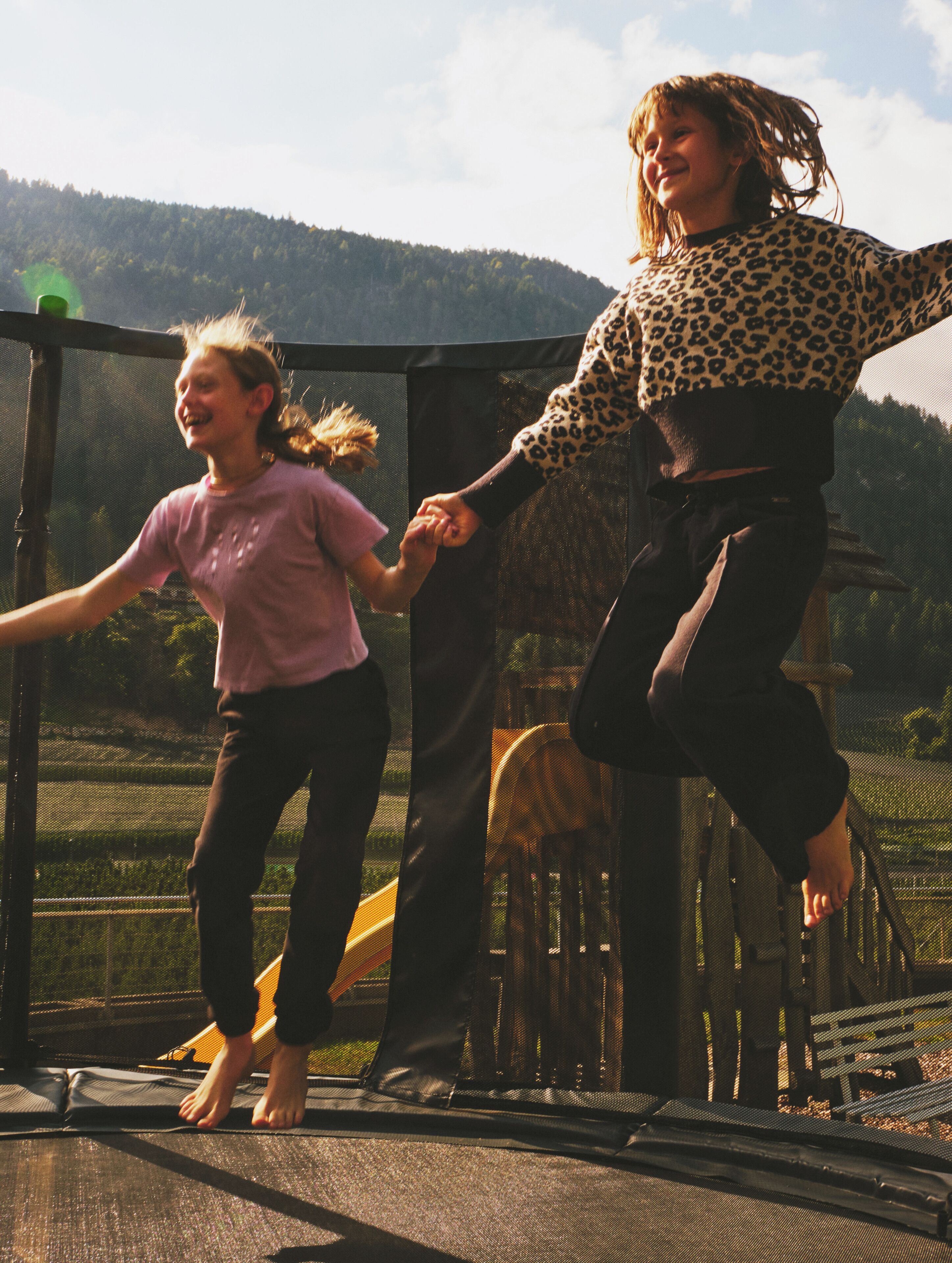 Two people jumping on a trampoline with a forested hill in the background.