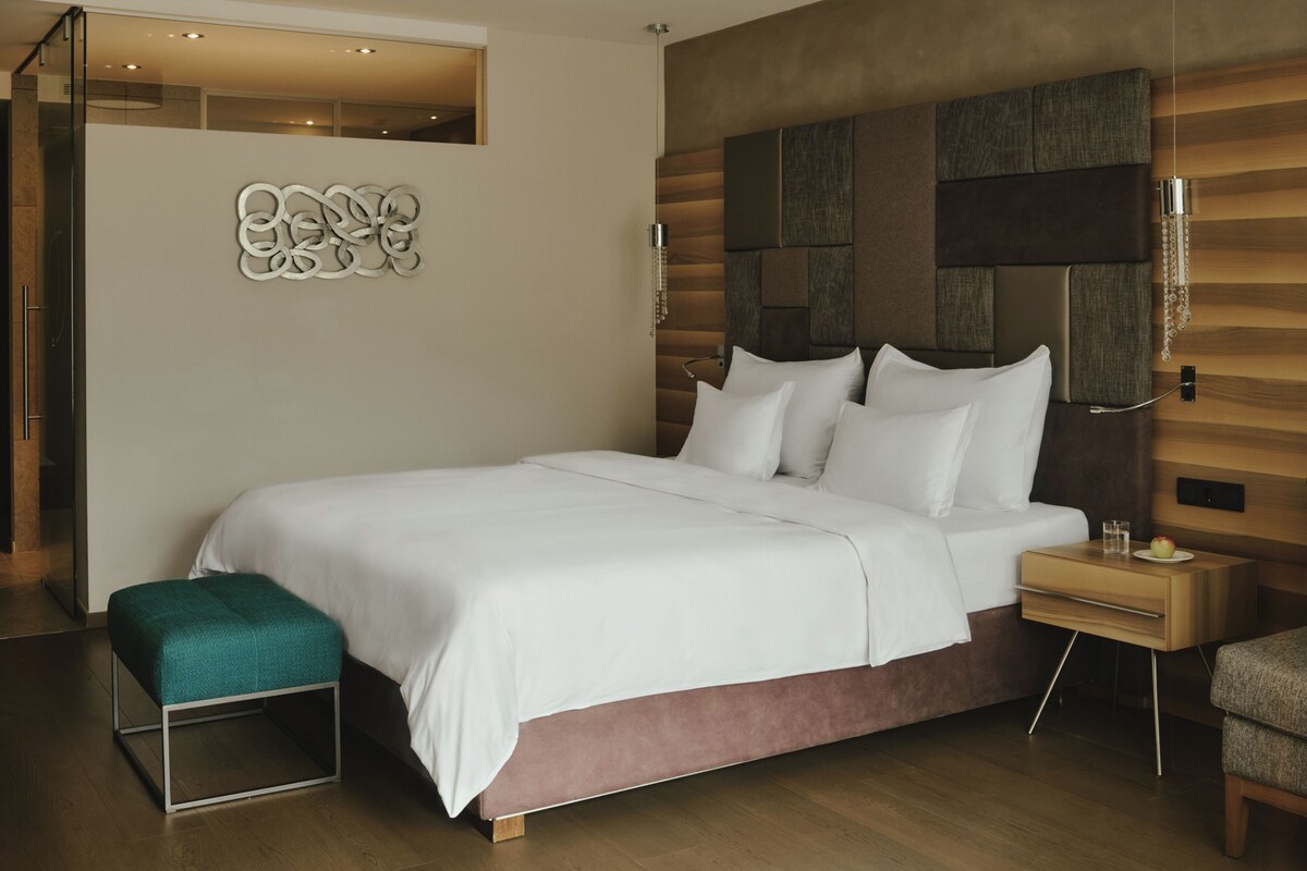 Quarto de hotel moderno com uma cama grande, uma mesinha de cabeceira e um pequeno banquinho.