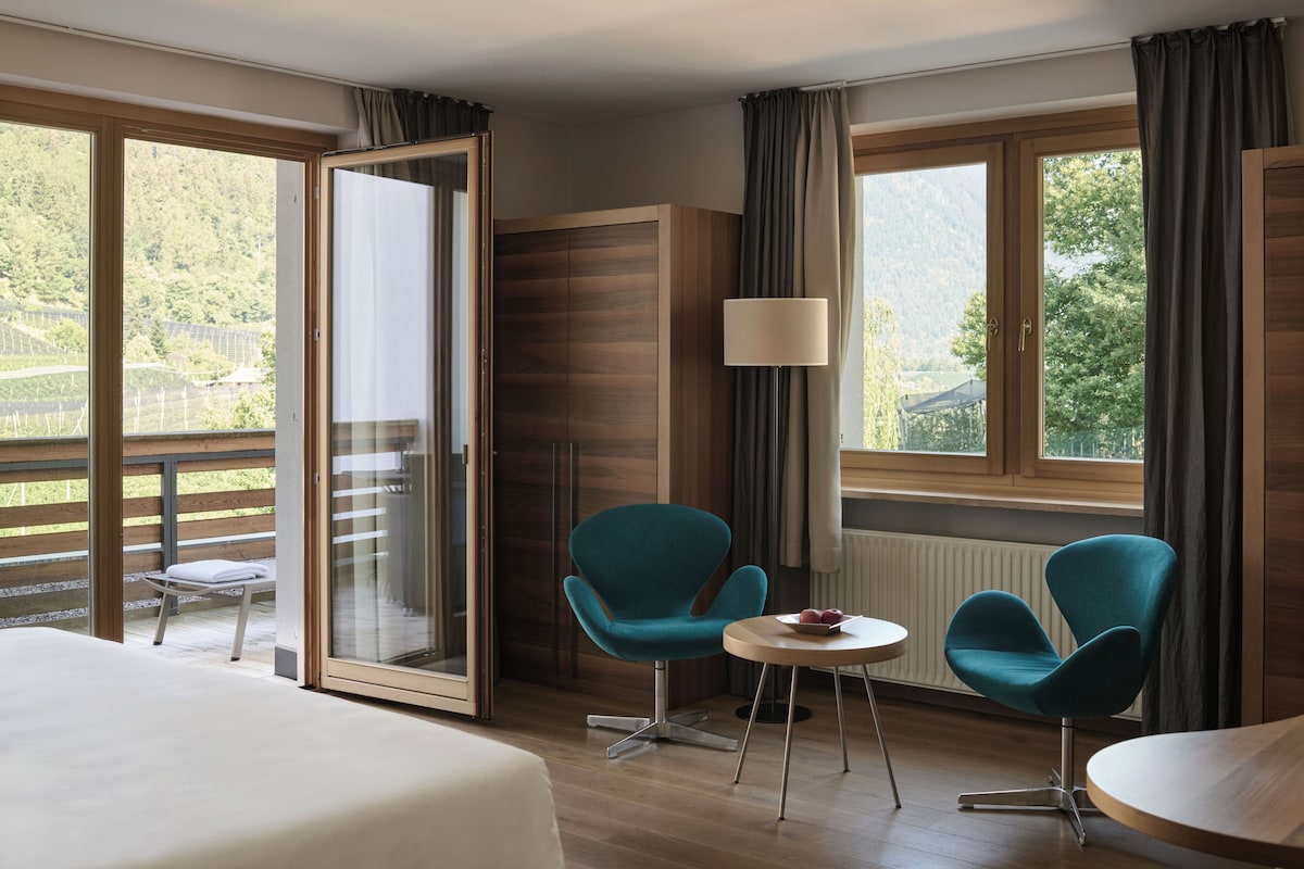 Quarto de hotel com uma cama, duas cadeiras, uma mesa, um abajur e vista para o exterior.