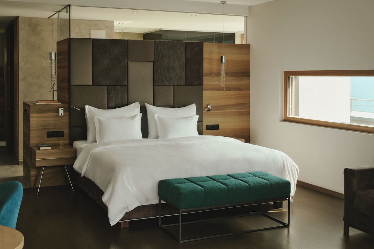 Quarto de hotel moderno com uma cama grande, um banco verde, uma mesa de cabeceira e uma janela com vista.
