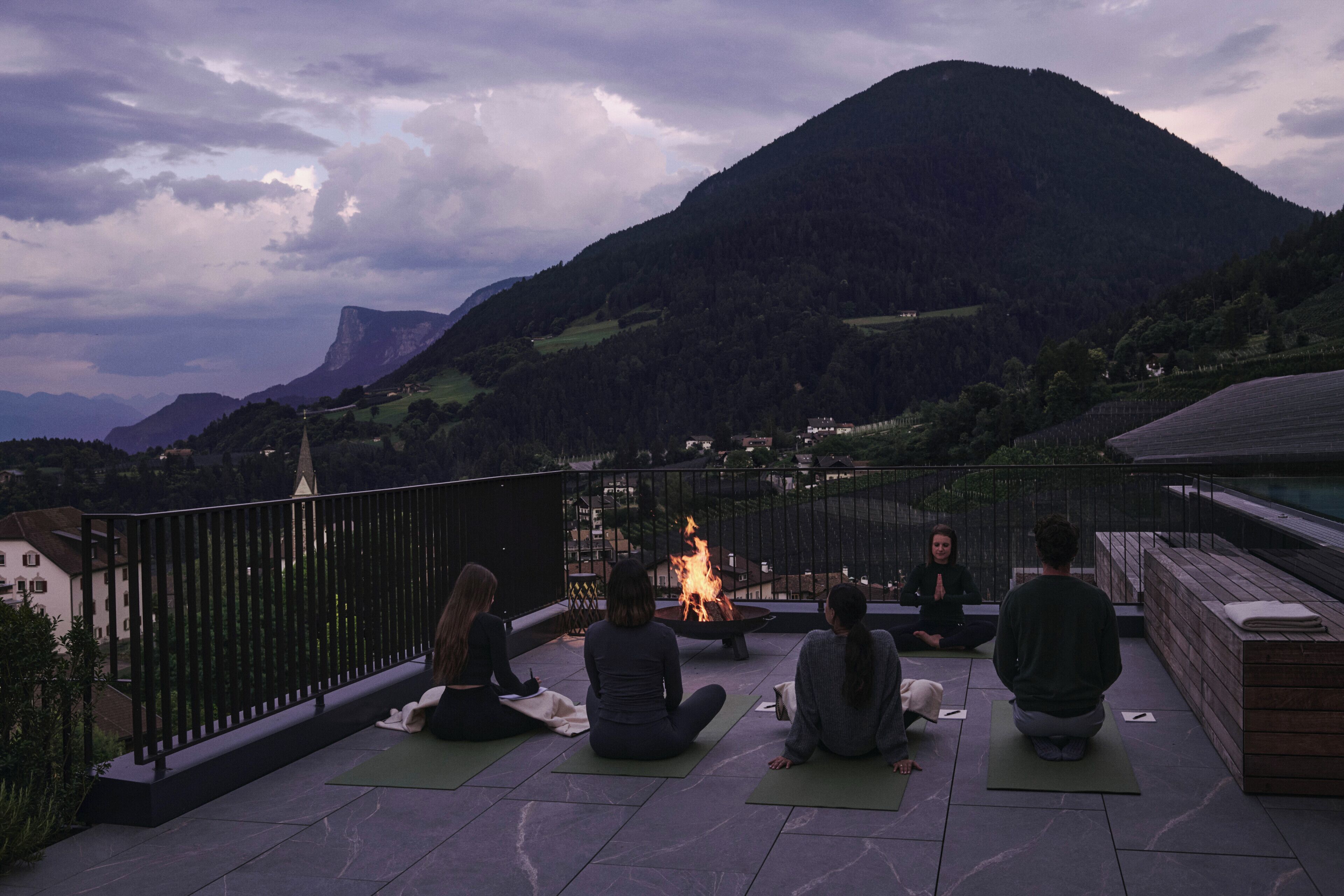Un gruppo di persone sedute su tappetini da yoga su una terrazza con un braciere, con vista su un paesaggio montuoso.
