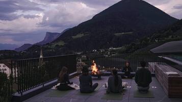 Un gruppo di persone sedute su tappetini da yoga su una terrazza con un braciere, con vista su un paesaggio montuoso.