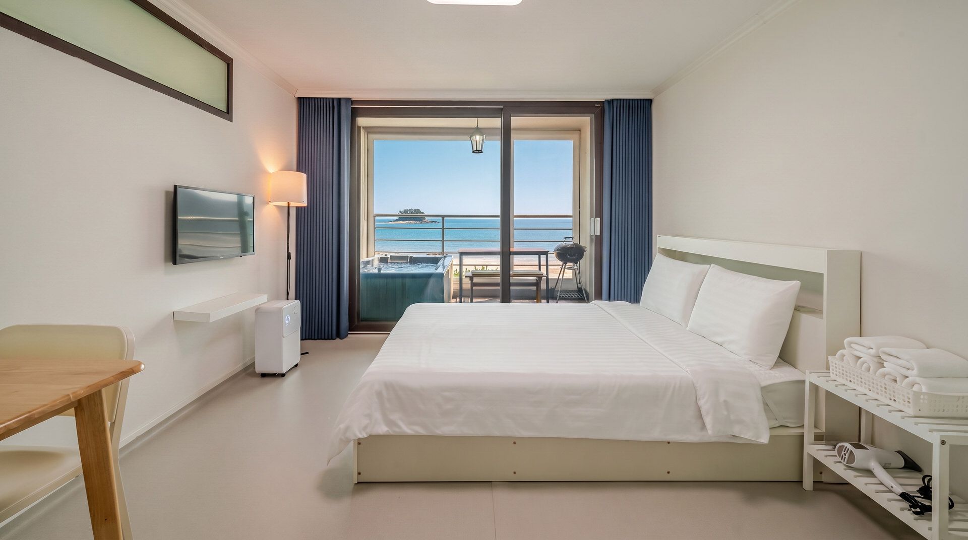 A hotel room with a bed, a desk, a TV, a heater, a table, a chair, a balcony with a view of the sea, and a towel rack.