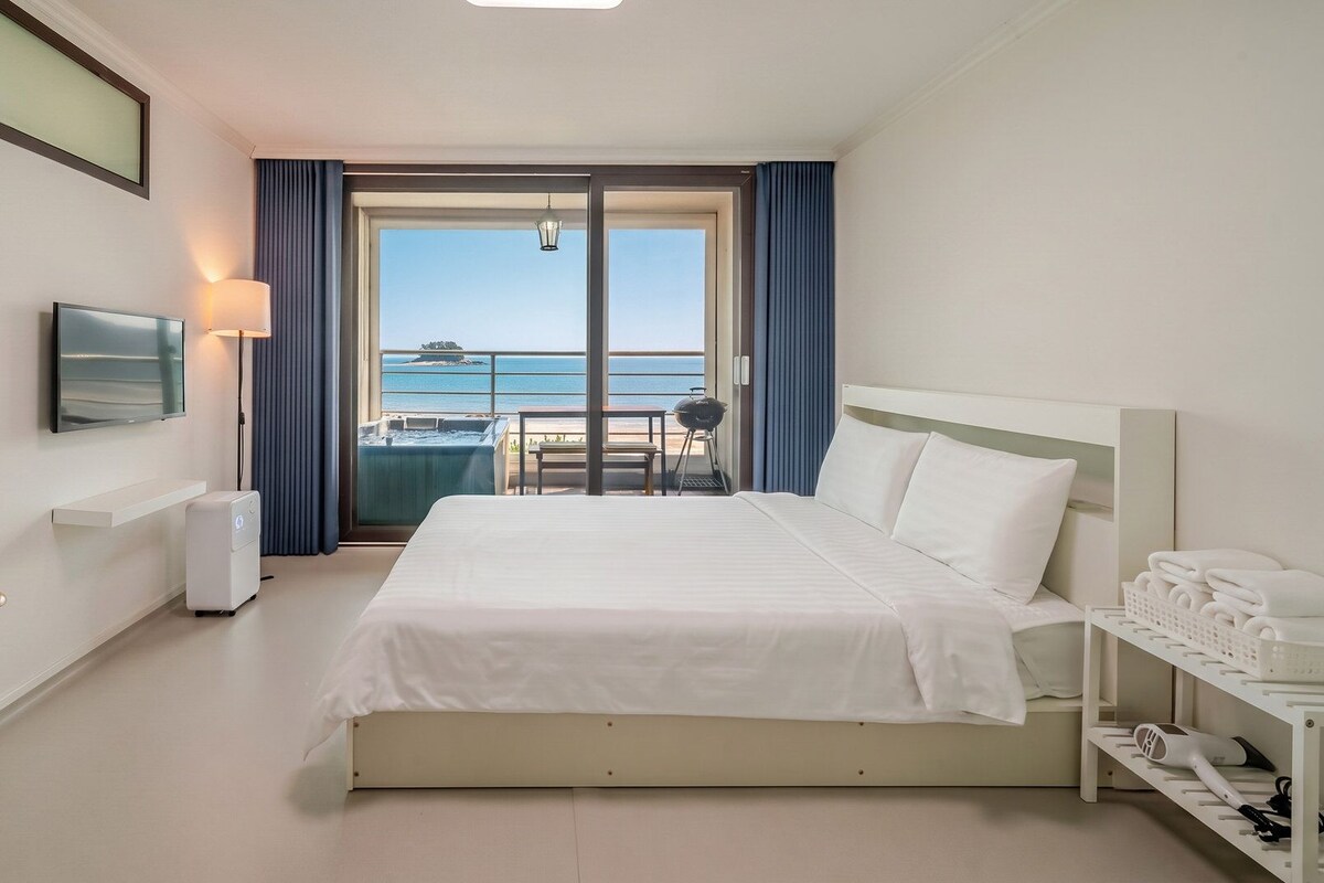 A hotel room with a bed, a desk, a TV, a heater, a table, a chair, a balcony with a view of the sea, and a towel rack.