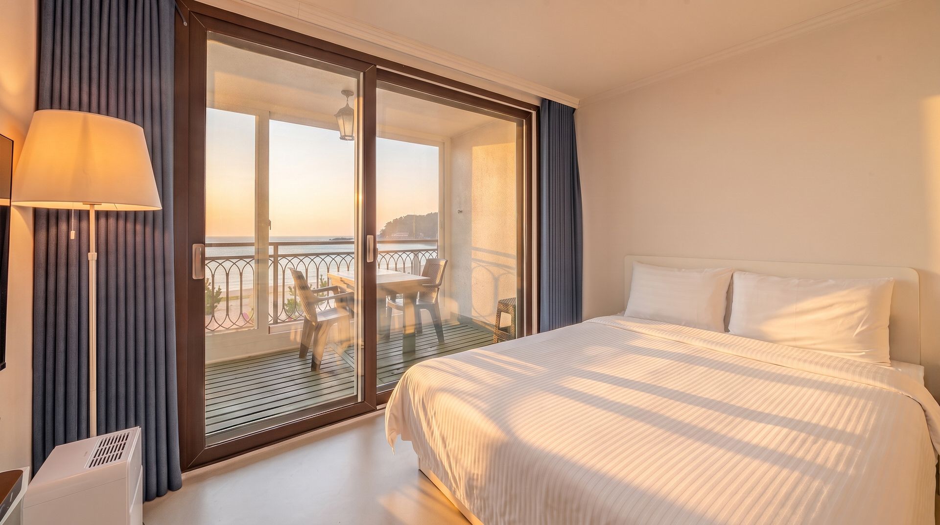 A hotel room with a bed, a desk with a lamp, a balcony with a table and chairs, and a view of the sea.