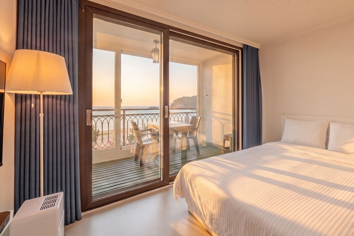 A hotel room with a bed, a desk with a lamp, a balcony with a table and chairs, and a view of the sea.