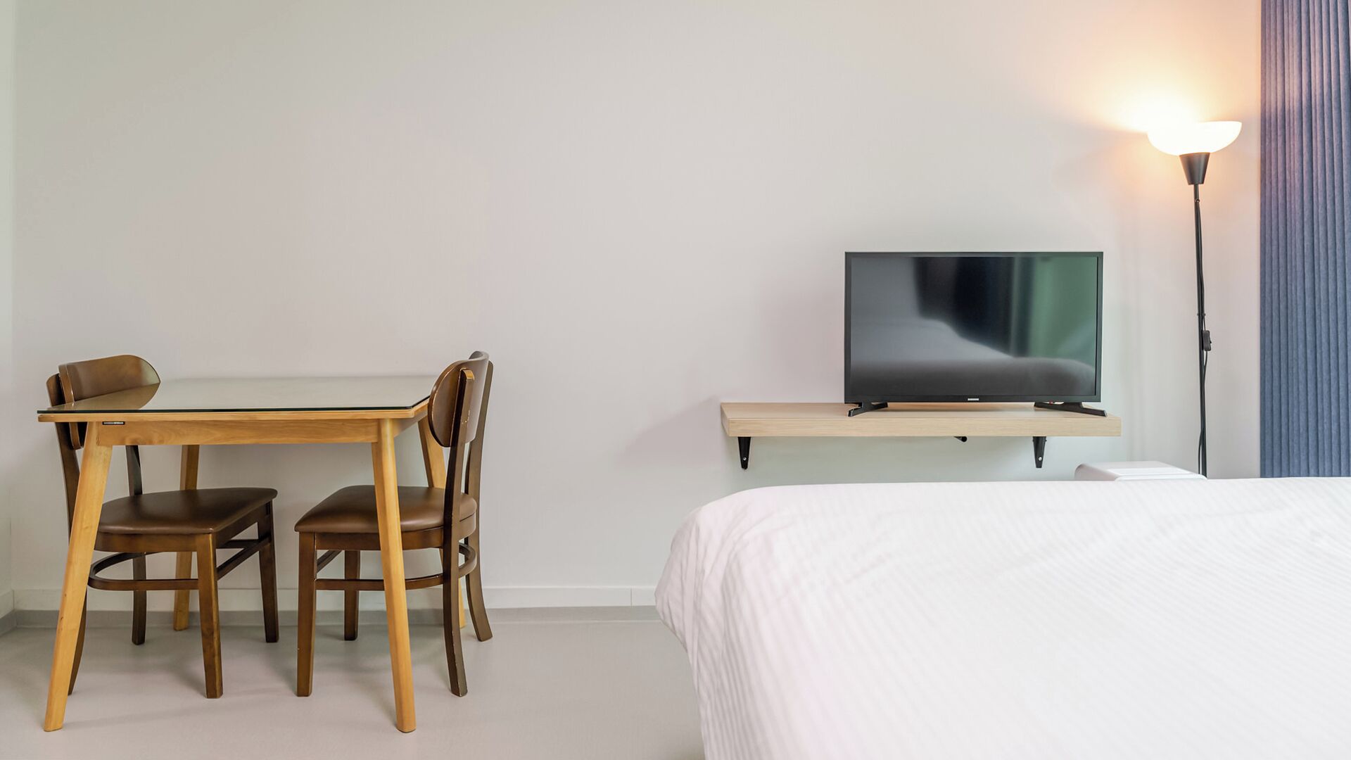 A hotel room with a bed, a TV on a stand, a desk with chairs, and a lamp.