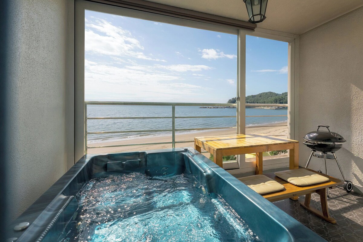A balcony with a hot tub, a table, and a grill, overlooking a beach and water.