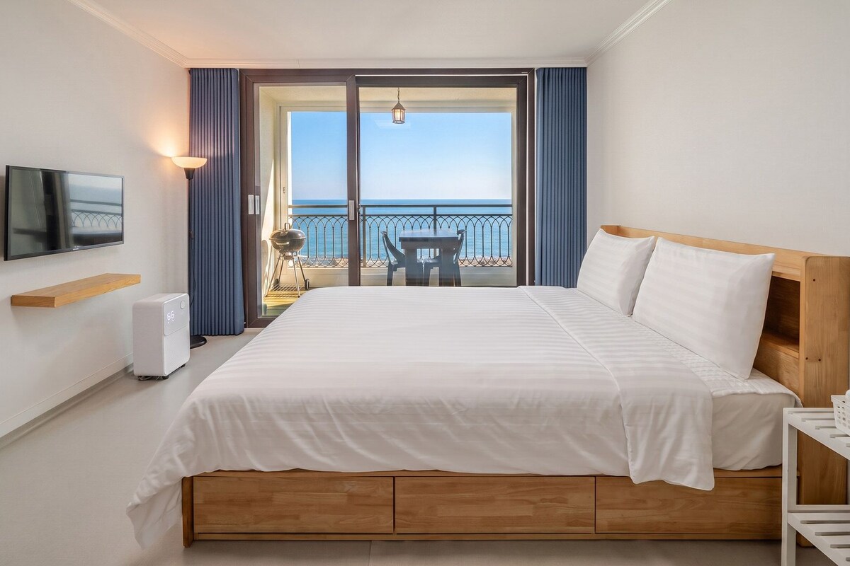 A hotel room with a bed, a TV, a heater, a table with chairs, and a view of the sea.