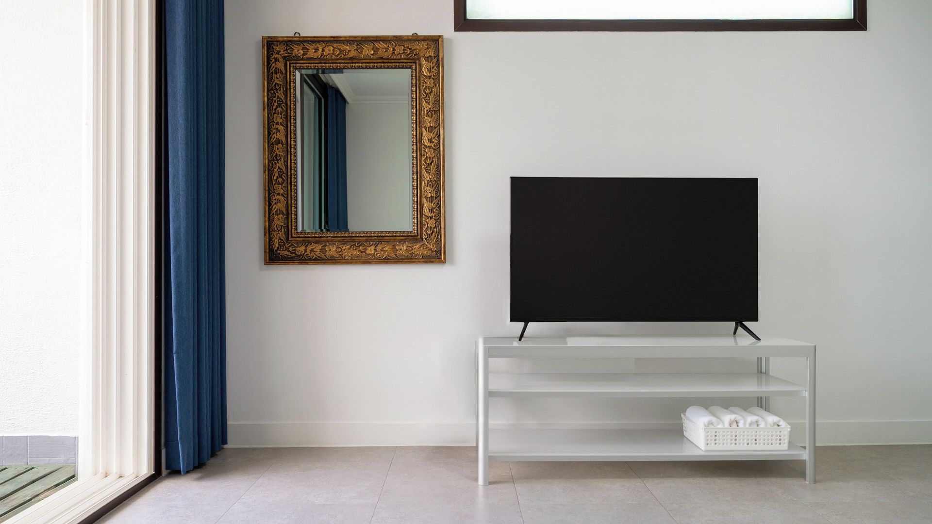 A room with a television on a white stand, a gold-framed mirror on the wall, and a window with blue curtains.